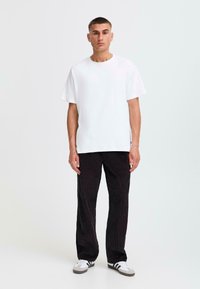 Weißes Baumwoll-T-Shirt mit kurzen Ärmeln, kombiniert mit schwarzen Cordhosen mit texturierten vertikalen Streifen, abgerundet mit schwarz-weißen Turnschuhen.