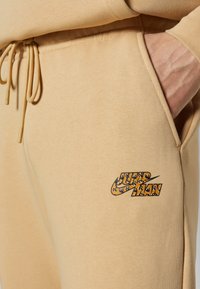 Beige Jogginghosen aus weichem Stoff mit einem Kordelzugbund und einem in Gelb und Schwarz gedruckten Nike-Logo oberhalb einer Tasche.