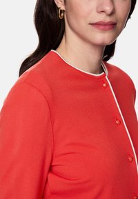Red knit top with a round neckline, contrasting white trim, and three button closures. Features a smooth texture and long sleeves.