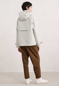 Vaaleanharmaa vedenpitävä takki hupulla, jossa on kerrostettu selkädesign ja sivunappikoristeet, yhdistettynä ruskeisiin corduroy-housuihin ja valkoisiin lenkkikenkiin.