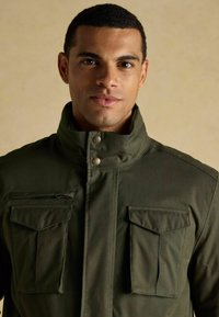 Olive o zielona kurtka z wysokim kołnierzem, dwiema kieszeniami na klatce piersiowej z klapami oraz kieszenią na zamek po lewej stronie. Gładka tekstura materiału.