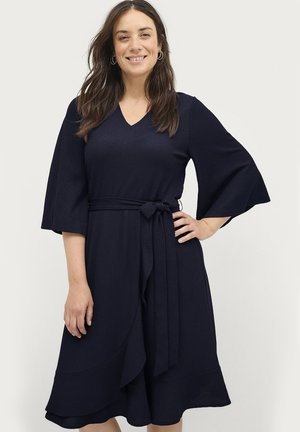 Kvinde med langt mørkt hår iført en marineblå kjole med bælte og brede ærmer, stående med den ene hånd på hoften og smile.