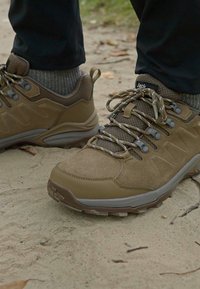Brune tursko laget av semsket skinn og nettingmaterialer, med gummisåle, teksturerte detaljer og kontrasterende lisser mot en sandaktig bakgrunn.