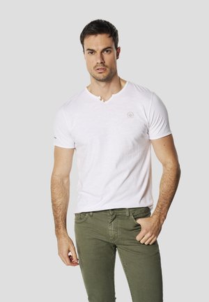 Camiseta blanca de algodón con cuello en V, con dos pequeños botones de madera en el escote. Combinada con jeans ajustados de color verde oliva.