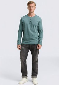 Langärmeliges tealfarbenes Henley-Shirt mit einer Drei-Knopf-Leiste, kombiniert mit dunkelgrauen Jeans und weißen Turnschuhen, zeigt eine lässige Passform.
