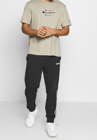 Béžové bavlněné tričko s logem Champion, spárované s černými tepláky s bílými bočními pruhy a logem Adidas, nošené s bílými teniskami.
