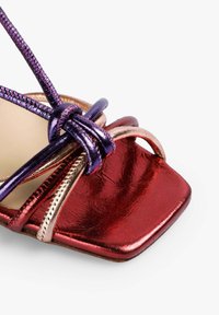 Hæl sandal med glinsende røde og metalliske lilla stropper, firkantet tådesign og intrikate knudede taljer. Glat tekstur hele vejen igennem.