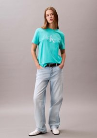 T-shirt di cotone turchese con logo bianco, abbinata a jeans in denim azzurro chiaro e scarpe bianche. Vestibilità comoda e stile casual.