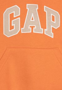 Oranžová mikina s nápisem "GAP" ve velkých, béžových, vyšitých písmenech. Obsahuje přední klokaní kapsu a strukturovanou látku.