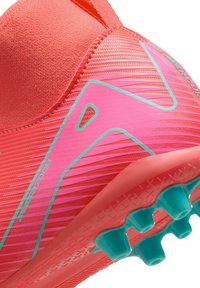Botas de fútbol de un vibrante color coral y rosa, con parte superior texturizada y acanalada, talón moldeado y tacos turquesa en la suela para ofrecer tracción.