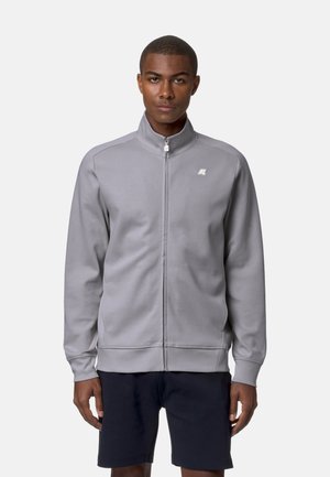 Uomo che indossa una giacca leggera grigio chiaro con una piccola logo sul petto e pantaloncini neri, in piedi davanti a uno sfondo chiaro e uniforme.