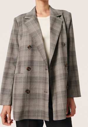 Femme portant un blazer croisé à carreaux gris avec des boutons marron sur un top blanc côtelé et un pantalon noir.