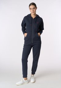 Felpa con zip di colore blu navy, con tasche frontali e dettagli rossi, abbinata a pantaloni jogger coordinati. Il materiale è morbido, con una texture liscia. Sneakers bianche completano l'aspetto.