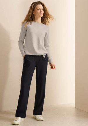 Maglietta grigia a maniche lunghe con scollatura a giro e nodo laterale. Pantaloni blu scuro con vestibilità ampia e tasche. Sneakers bianche completano l'outfit.