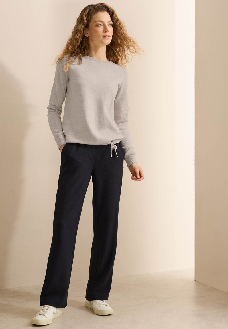 Maglietta grigia a maniche lunghe con scollatura a giro e nodo laterale. Pantaloni blu scuro con vestibilità ampia e tasche. Sneakers bianche completano l'outfit.