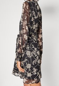 Kvinna i en svart genomskinlig klänning med beige blommönster och repmönster, långa ärmar och knytning i midjan.