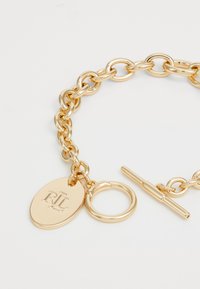 Zlatý náramok s oválnym príveskom "RL" a otočným zapínaním na bielom pozadí.