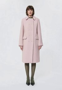 Γυναίκα που φοράει ένα ροζ παλτό μέχρι το γόνατο, διπλής όψης, με τσέπες παράθυρο, ελιά καλσόν και μαύρες μυτερές γόβες, στέκεται μπροστά σε απλό φόντο.
