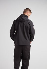 Giacca nera impermeabile con cappuccio, polsini elastici e logo sulla manica, abbinata a pantaloni neri e uno sfondo semplice.
