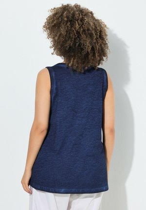 Person mit lockigem Haar, die ein ärmelloses dunkelblaues Oberteil und weiße Hose trägt, steht mit dem Gesicht zu einer hellgrauen Wand.