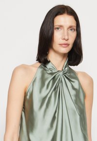 Matt zöld, nyakán csavart masni részlettel rendelkező halter top, amely sima, fényes textúrával és laza eséssel rendelkezik.