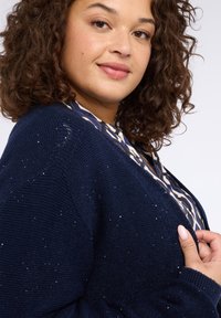Cardigan lavorato a maglia blu navy con dettagli a scintillio discreto. Presenta una vestibilità ampia, polsini a costine e una profonda scollatura a V, offrendo un aspetto strutturato.