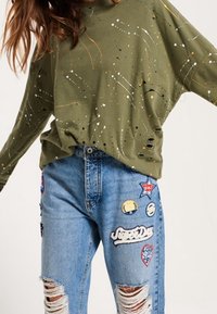 Sudadera oversize de color verde oliva con patrón de salpicaduras de pintura. Jeans desgastados de un azul claro con parches coloridos variados y desgarros.