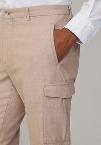 Beige cargobyxor i lätt tyg med sidoficka och detaljer vid sömmarna. Matchas med en vit skjorta med krage.