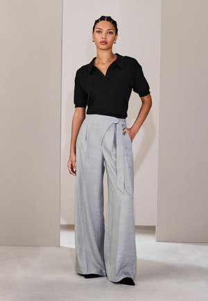 Kvinde står iført en sort kortærmet skjorte med krave og grå høj-taljede, vidbenede bukser med bånd i siden.