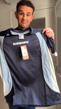 Άνδρας κρατάει μπλούζα αθλητική Umbro σε σκούρο μπλε χρώμα με ετικέτες σε ένα δωμάτιο με πόρτα και καθρέφτη πίσω του.