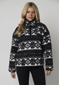 Donna che indossa un pullover di pile a maniche lunghe con un motivo in bianco e nero e una zip a un quarto, abbinato a leggings neri, mentre posa davanti a uno sfondo neutro.
