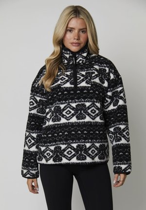 Mujer vestida con un pulóver de forro polar de patrón en blanco y negro con media cremallera, combinado con leggings negros, posando contra un fondo plano.