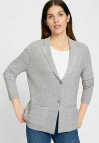 Grauer gerippter Cardigan mit einem Schalkragen, zwei vorderen Taschen und einem Knopfverschluss. Über einem weißen Langarmshirt getragen.