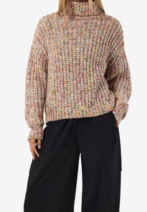 Vrouw draagt een dikke, meerkleurige coltrui en losse zwarte cargobroek met zakken aan de zijkant, hand in de zak.