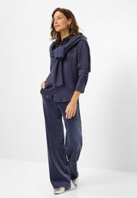 Hemd in Marineblau mit aufgerollten Ärmeln, kombiniert mit weit geschnittenen Marineblau-Hosen. Ein gestrickter Schal liegt lässig über den Schultern und wird von silbernen Schuhen ergänzt.