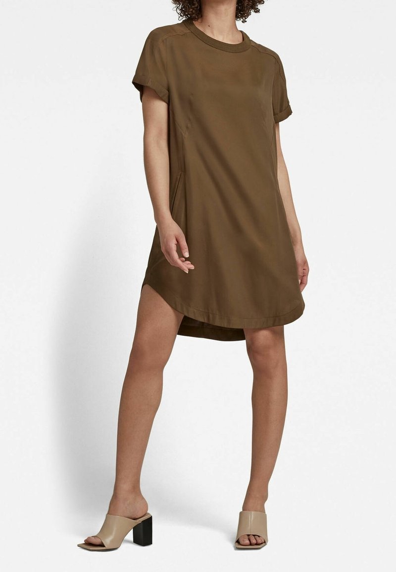 Femme portant une robe marron à manches courtes, ample, avec des poches latérales et des sandales ouvertes à talons beiges, debout devant un fond blanc.
