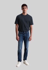 Donkergrijze katoenen T-shirt gecombineerd met blauwe spijkerbroek met vervagingdetails en witte sneakers. Model staat tegen een neutrale grijze achtergrond.