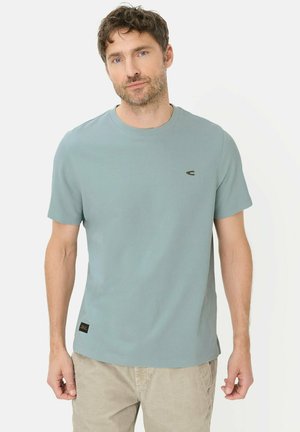 Man die een lichtblauw T-shirt met ronde hals en korte mouwen draagt en beige broek, staand tegen een effen lichte achtergrond.