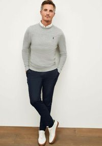 Pull gris avec texture côtelée, chemise blanche à col en dessous, pantalon marine et baskets blanches avec semelles marron. Détail de logo subtil sur le pull.