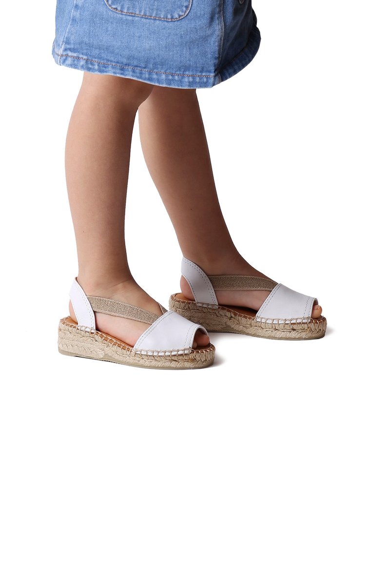 Enfant portant des sandales blanches à bout ouvert avec des brides élastiques beiges et des semelles compensées tissées, debout dans une jupe en jean.