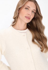 Młoda kobieta z długimi falującymi blond włosami, ubrana w kremowy, puszysty kardigan, pozująca na tle białego tła.