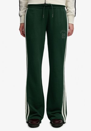 Mørkegrønne vidde joggingbukser med hvide siderstriber, snøre i taljen og "Superdry 89" tryk på venstre lår, brugt med mørke sko.