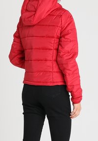 Giacca rossa invernale gonfia con cappuccio, caratterizzata da cuciture orizzontali, polsini elasticizzati e un design cropped, abbinata a pantaloni neri aderenti.