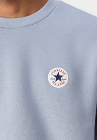 Lys blå sweatshirt med rund halslinning. Har et rundt merke med "Converse All Star"-tekst og et stjernedesign i blått og rødt.