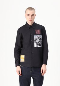 Black button-up shirt with various graphic patches, including a textured red square, a black and white illustration, and a colorful label.