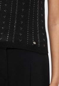 Črna pletena majica z navpičnimi vrstami majhnih sequinsov. Rebasta tekstura s srebrno logotipno značko na robu. Oblečena s črnimi hlačami.