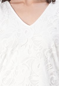 Haut en dentelle blanche avec un col en V, présentant des motifs floraux complexes et une finition texturée, offrant une apparence légère et respirante.