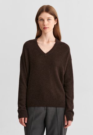 Jeune femme à la peau claire et aux cheveux bruns raides, portant un pull à col en V brun foncé et un pantalon gris, debout devant un fond uni.