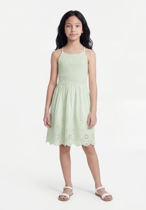 Κορίτσι που στέκεται με μακριά σκούρα μαλλιά, φοράει αμάνικο ανοιχτοπράσινο φόρεμα με τρυπητά σχέδια στο τελείωμα και λευκά σανδάλια σε λευκό φόντο.