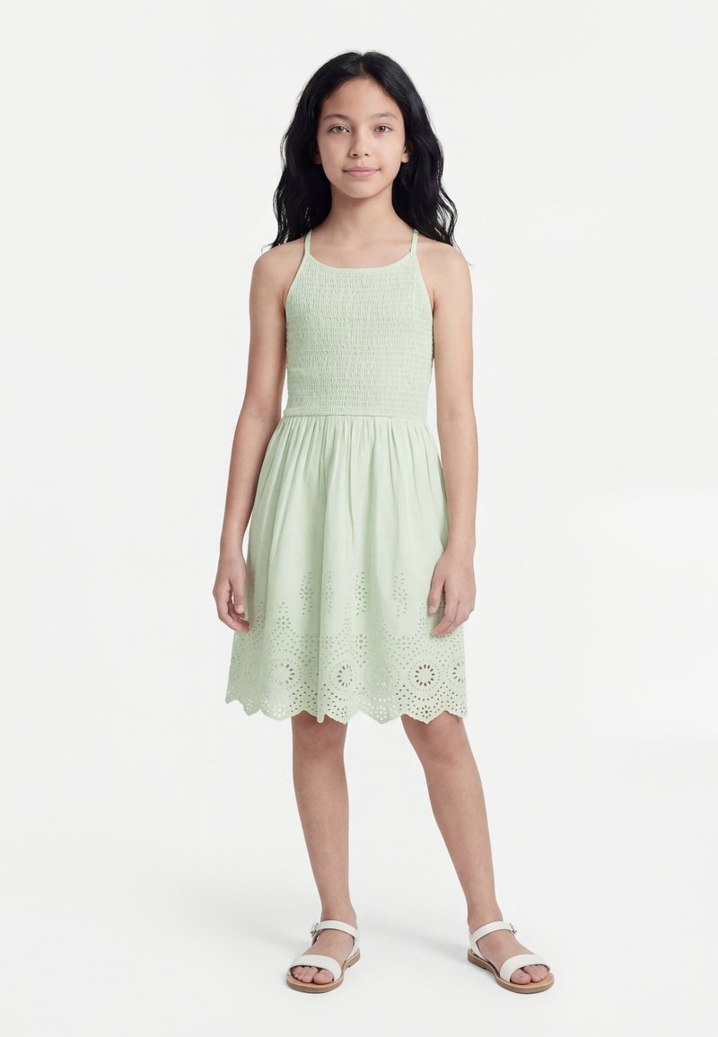 Κορίτσι που στέκεται με μακριά σκούρα μαλλιά, φοράει αμάνικο ανοιχτοπράσινο φόρεμα με τρυπητά σχέδια στο τελείωμα και λευκά σανδάλια σε λευκό φόντο.